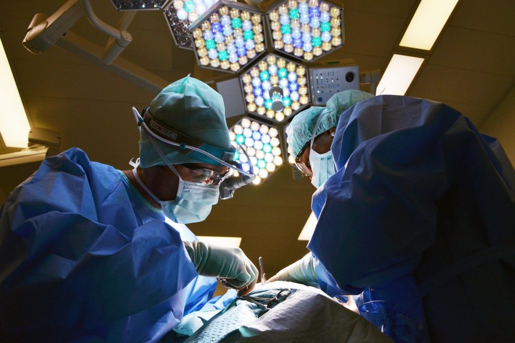 Two surgeons, clad in blue surgical gowns and masks, perform surgery under bright operating room lights. Meticulously focused on the task at hand, they work amidst a sea of medical equipment—ever mindful of the precision required to prevent any hint of medical negligence.