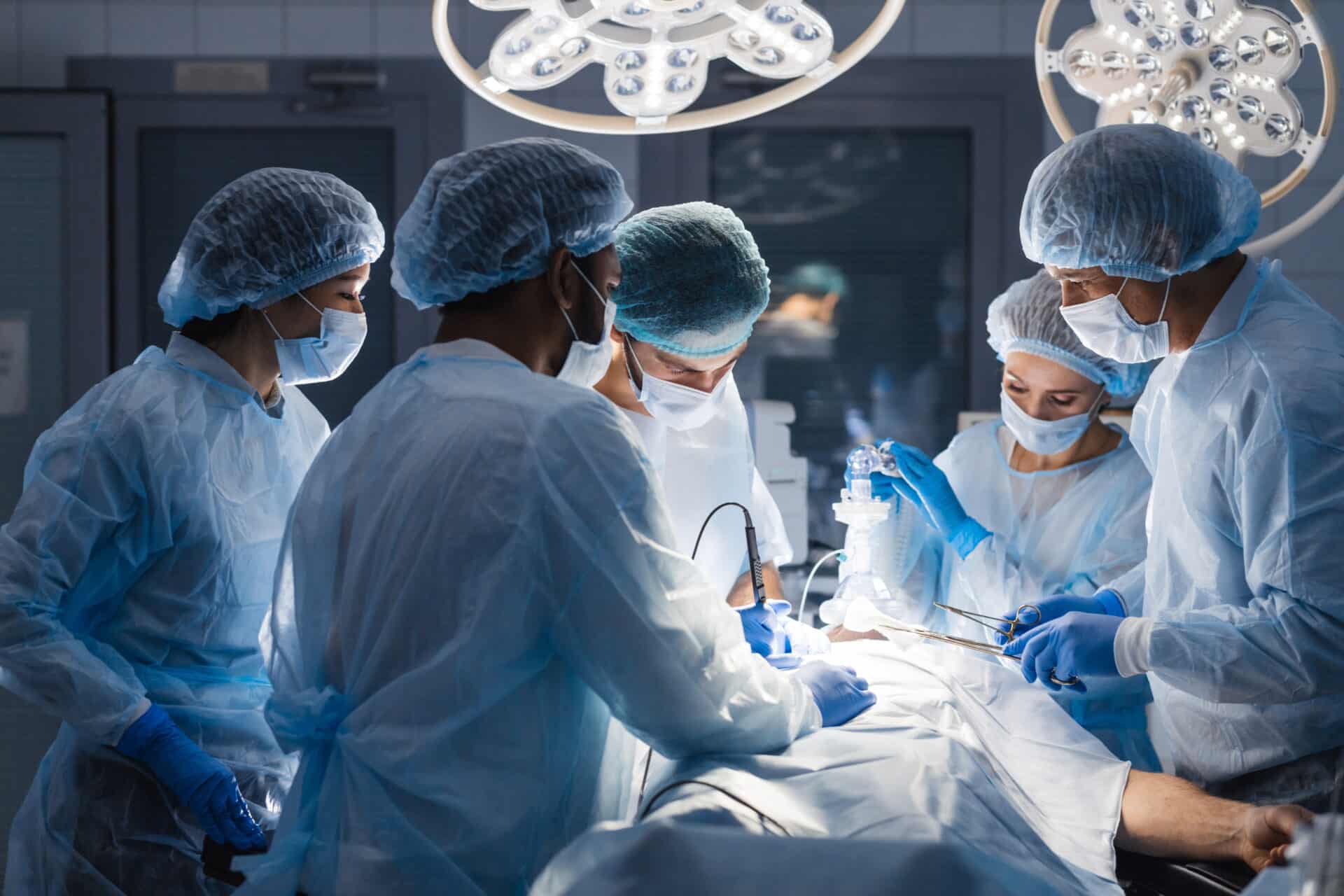 A team of surgeons in blue scrubs and masks performs a surgical procedure under bright overhead lights. They are focused, with various surgical tools and equipment visible around them in a sterile operating room environment.