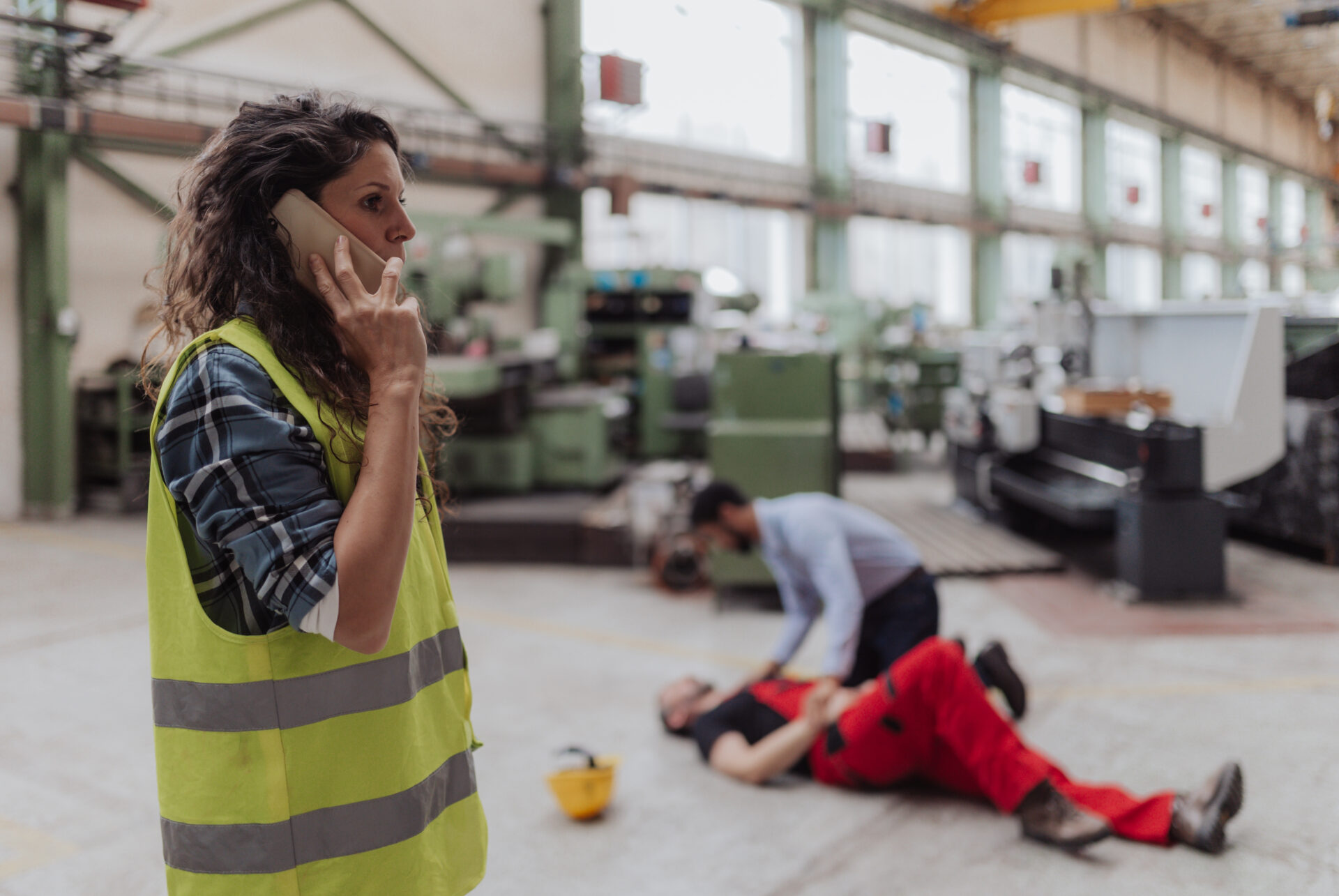A woman in a yellow safety vest is on the phone in a factory. In the background, a person kneels beside a person in red overalls lying on the floor, appearing to assist them. Industrial equipment is visible around them.