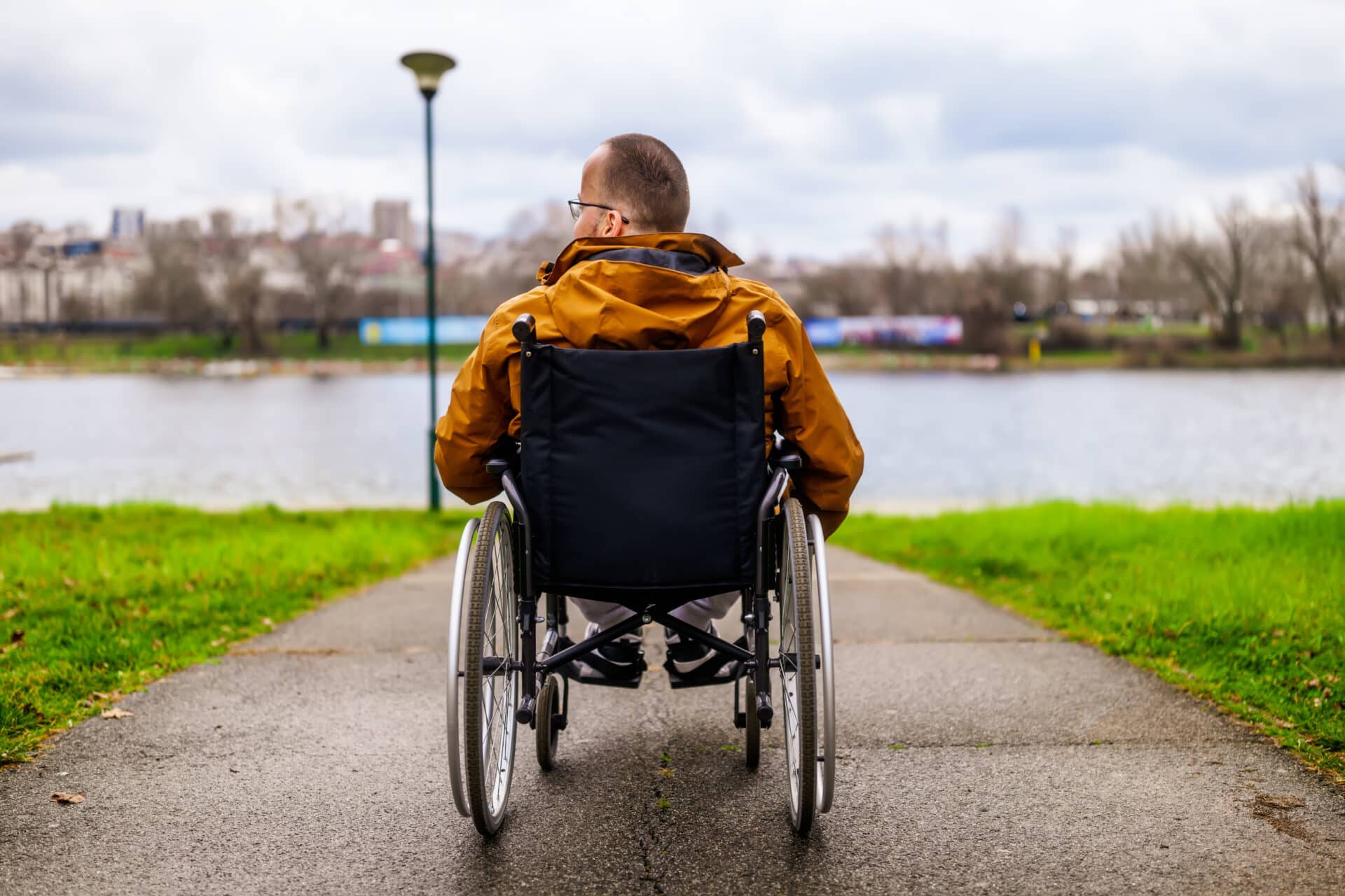 A person in a wheelchair faces a lake on a paved path. They wear a brown jacket and glasses. The overcast sky and bare trees suggest a chilly day. A lamppost is nearby, and a distant city skyline is visible in the background.