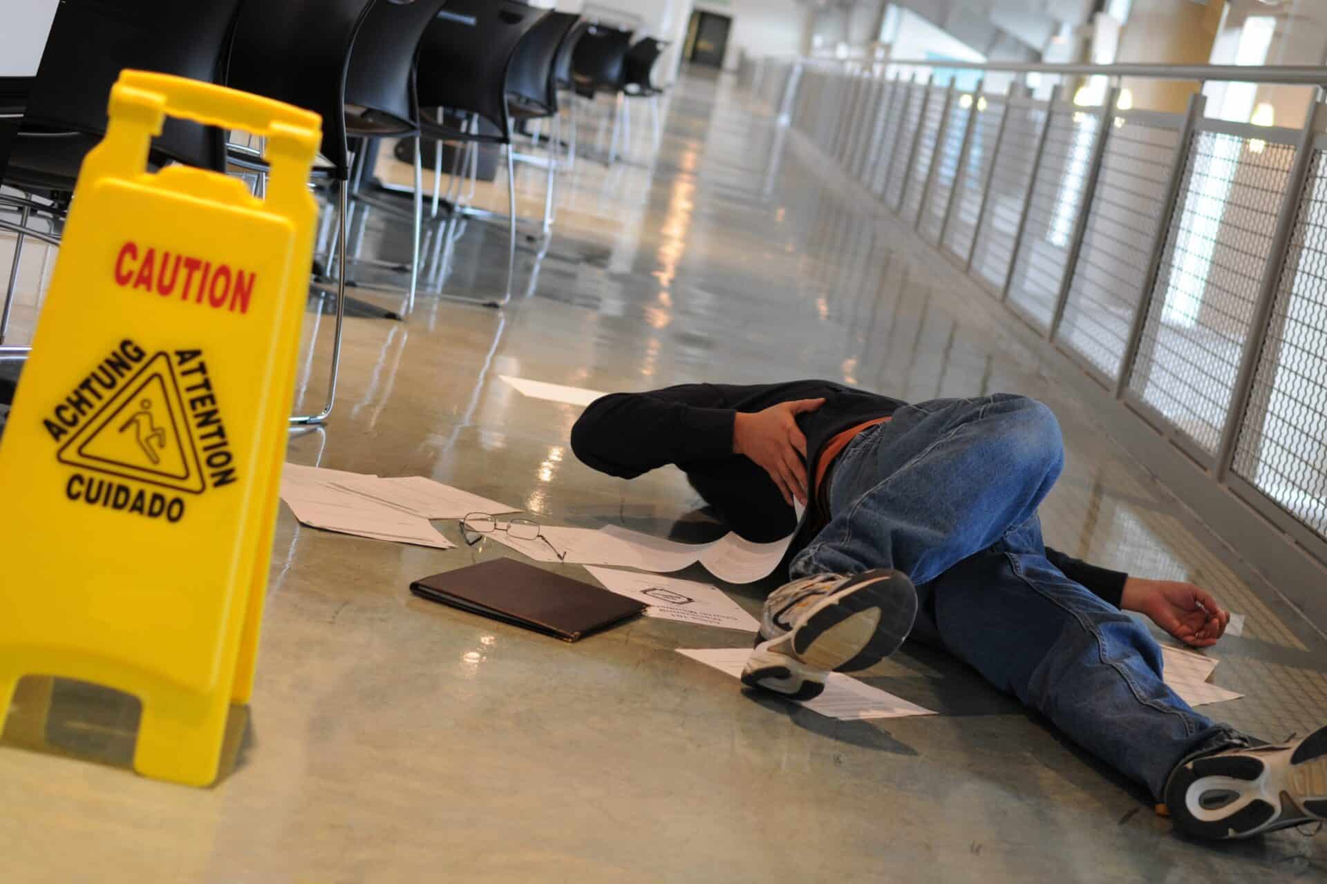 A person lies on the floor of a hallway next to scattered papers and a folder, appearing to have fallen. A yellow caution sign is visible nearby, indicating a wet surface. Chairs and a glass railing are in the background.